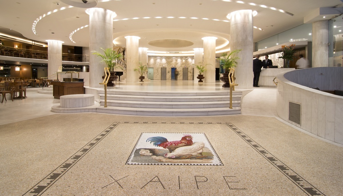 A picture of Hotel Titania's lobby showing a staircase surrounded by white columns, a check-in desk, and tiled floor with a mosaic that says 'Xaipe' in Greek ('Rejoice').