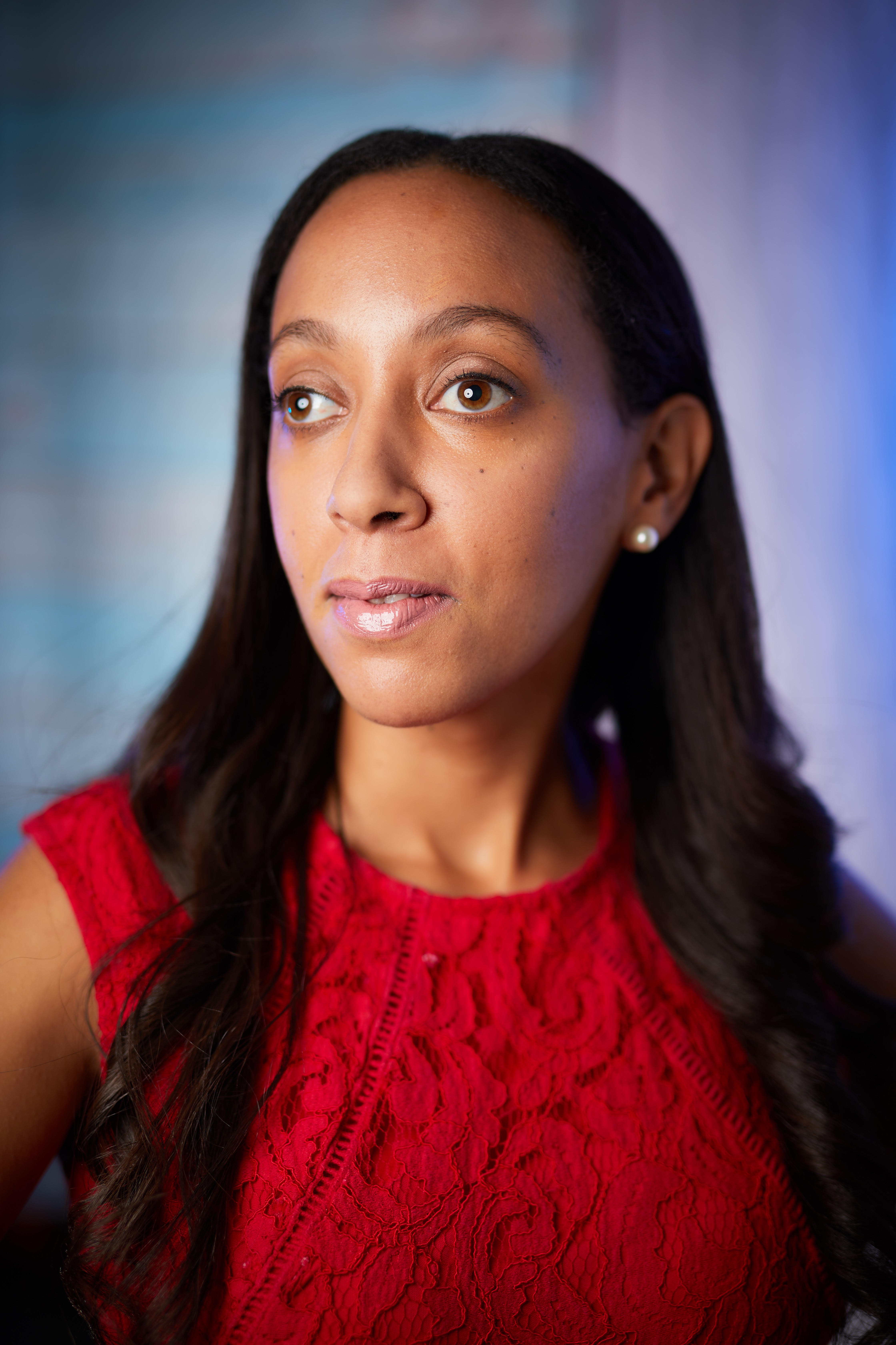 A 30-ish, Black woman with long dark hair and hazel eyes, wearing a textured red top and a thoughtful expression. The background has shades of blue.