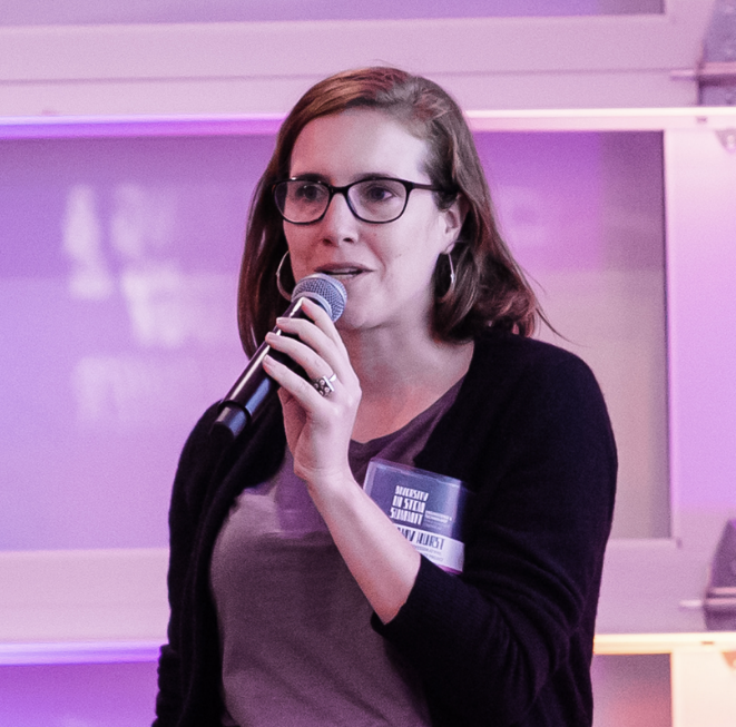 Headshot of Amy Hurst from the shoulders up holding a microphone and talking into it. Amy is a white woman with shoulder-length brown hair and glasses. 