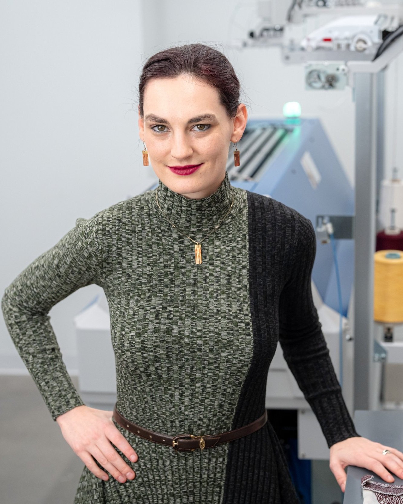 A photo of Megan Hofmann, a White woman with purple hair in a tight bun. Shes wearing a green and black knit dress and bright lipstick. She leans on a lab table in front of a knitting machine.