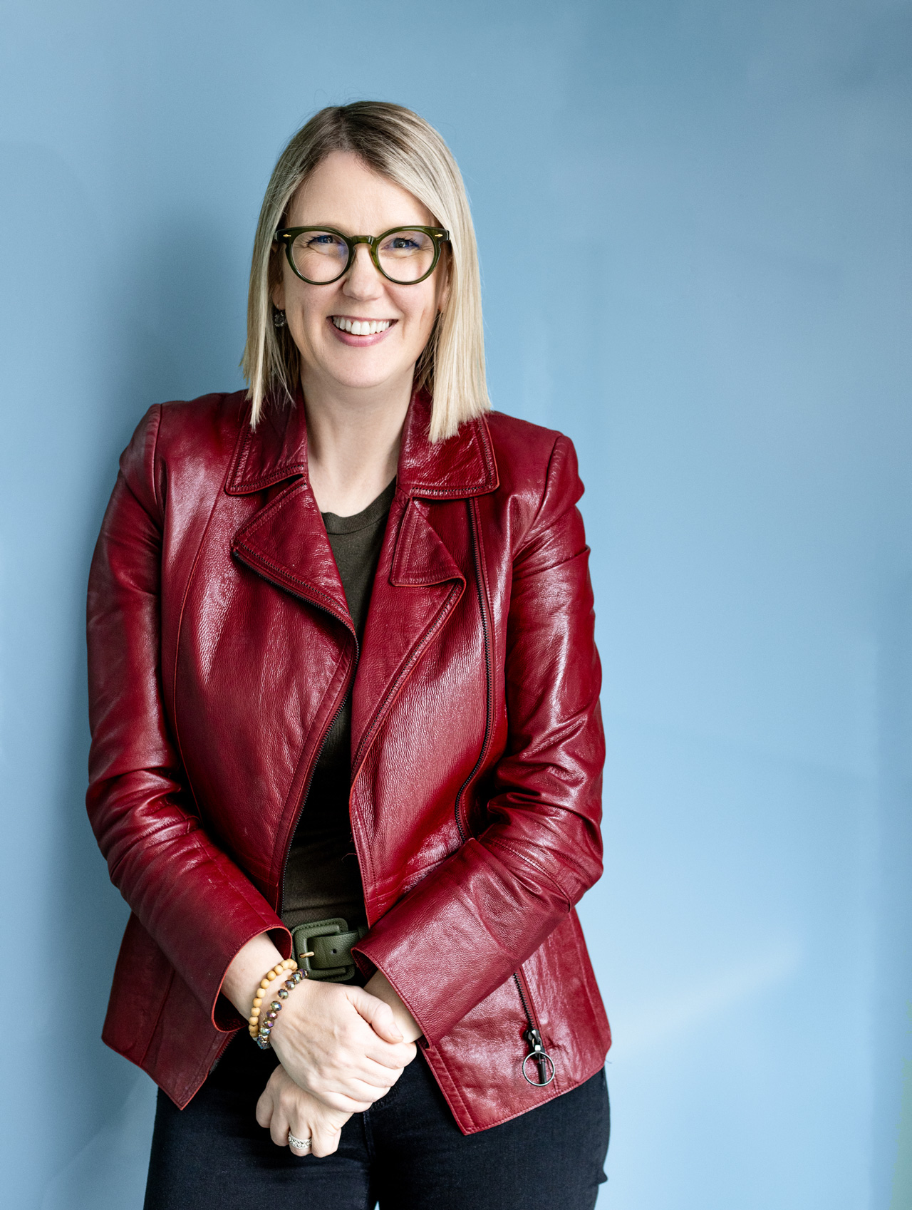 Jenny Lay-Flurrie, a woman with straight blonde hair and green-framed glasses, smiling while wearing a red leather jacket over a dark top and black pants, standing against a light blue background with her hands clasped in front.