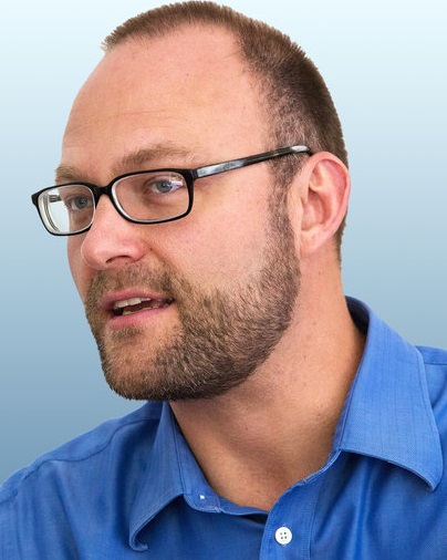 Jacob O. Wobbrock, a man with short brown hair, a beard, and glasses, wearing a blue collared shirt and looking slightly to the side against a light blue background.