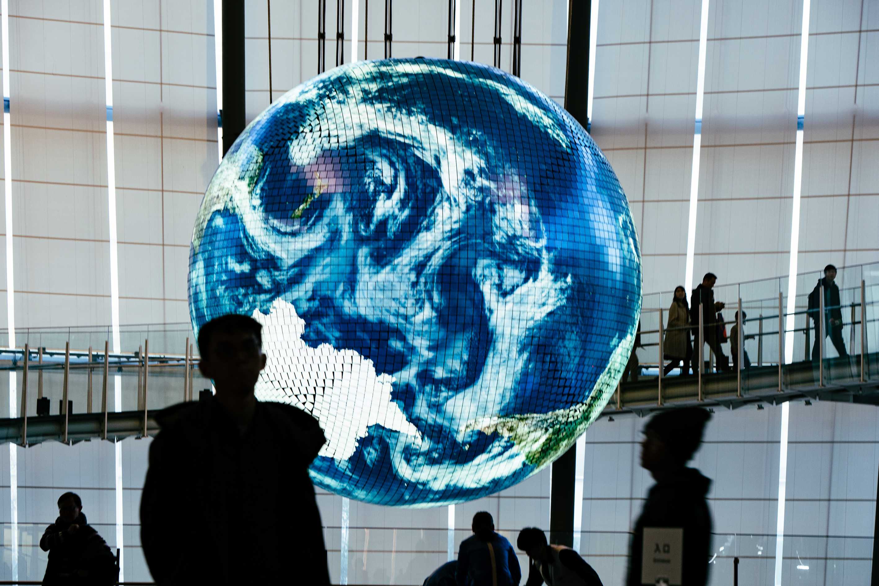 Inside Miraikan: the Geo-Cosmos, a large glowing globe showing the Earth and swirling clouds, hangs in a tall white atrium while visitors, seen in silhouette, walk below and along a glass-railed ramp.