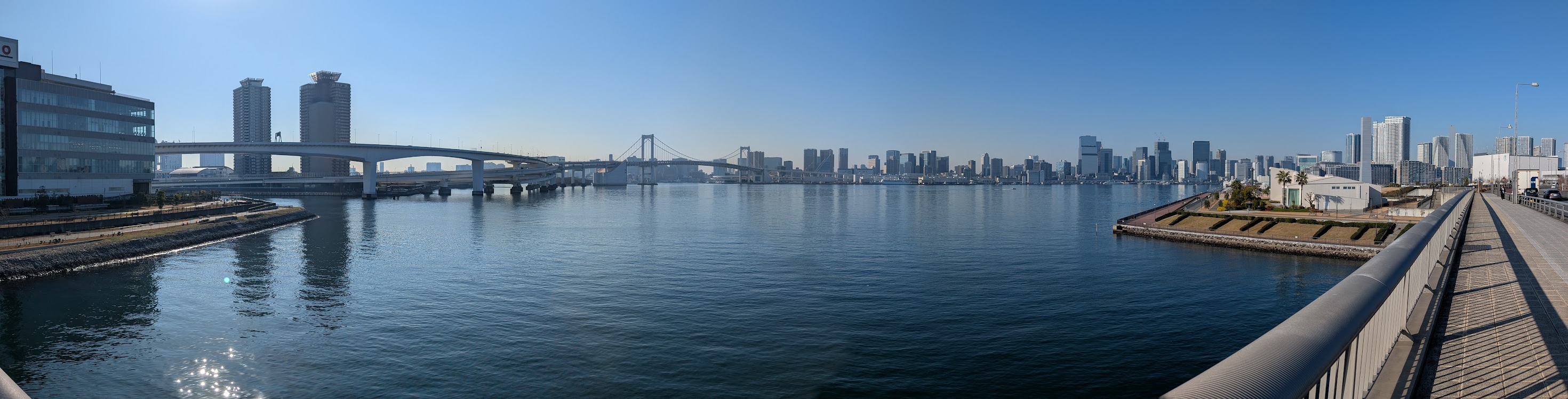 Panoramic view of Tokyo Bay on a clear day: the Rainbow Bridge spans the water in the centre, with the Tokyo city skyline behind it, a curving elevated highway and two tall towers on the left, and a waterfront promenade with palm trees on the right.